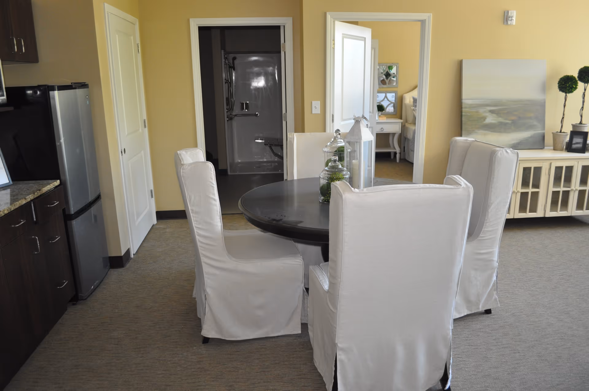 Interior view of a room in an assisted living facility showing a round dining table with four white slipcovered chairs. To the left is a kitchenette with dark cabinets and a small refrigerator. In the background, there are two open doorways, one leading to a bathroom with a shower and the other to a bedroom with a white nightstand and bed. On the right side, there is a white cabinet with decorative items including a painting and small plants.