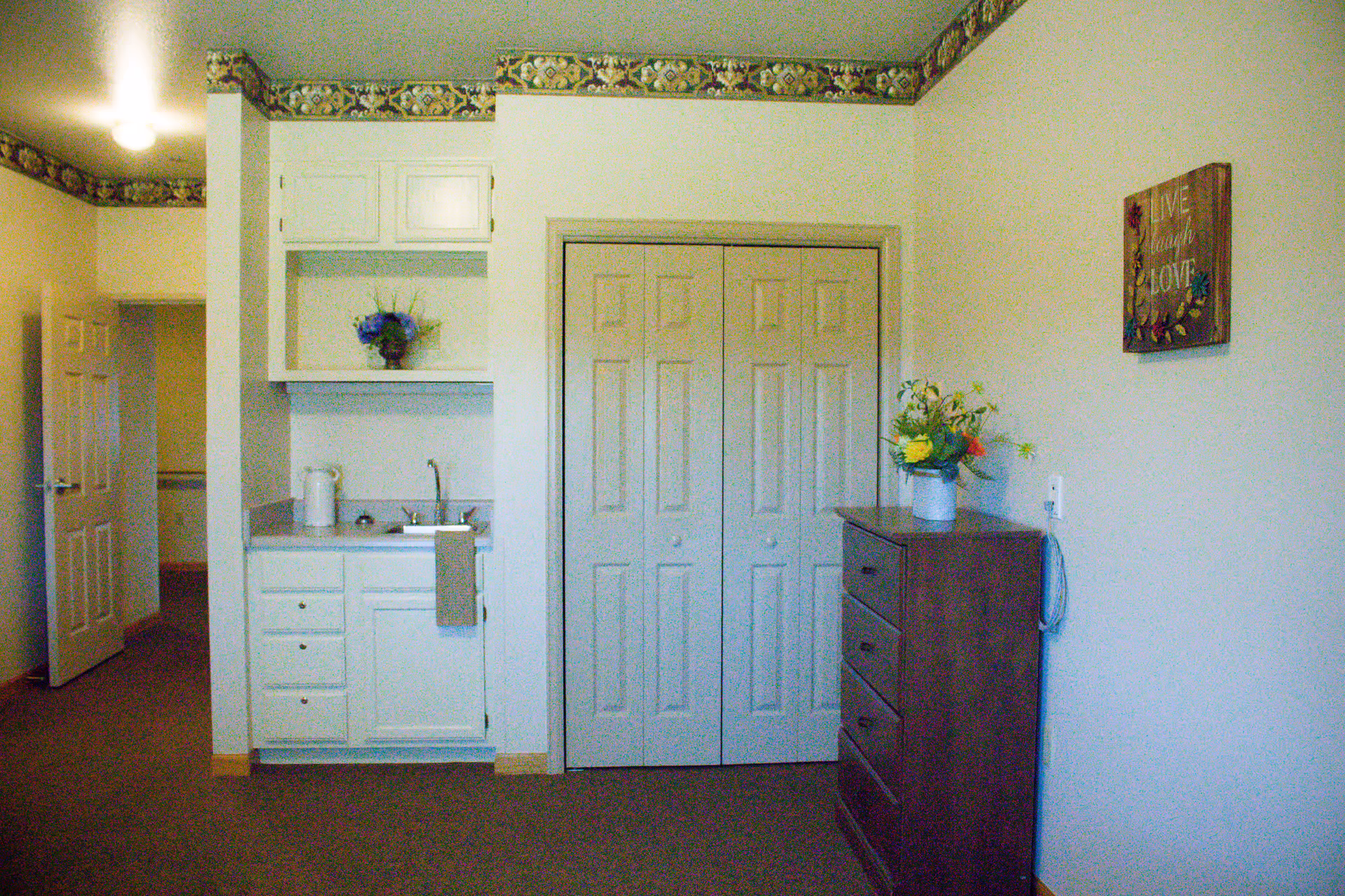 Small assisted living studio room with a kitchenette, double-door closet, and a dresser topped with flowers.