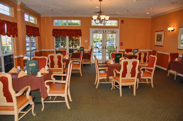 A dining room with several tables covered in brown tablecloths, each set with napkins, cups, and menus. The chairs have wooden frames with red cushioned backs and seats. Large windows with red valances let in natural light, and a chandelier hangs from the ceiling. Double glass doors lead outside.