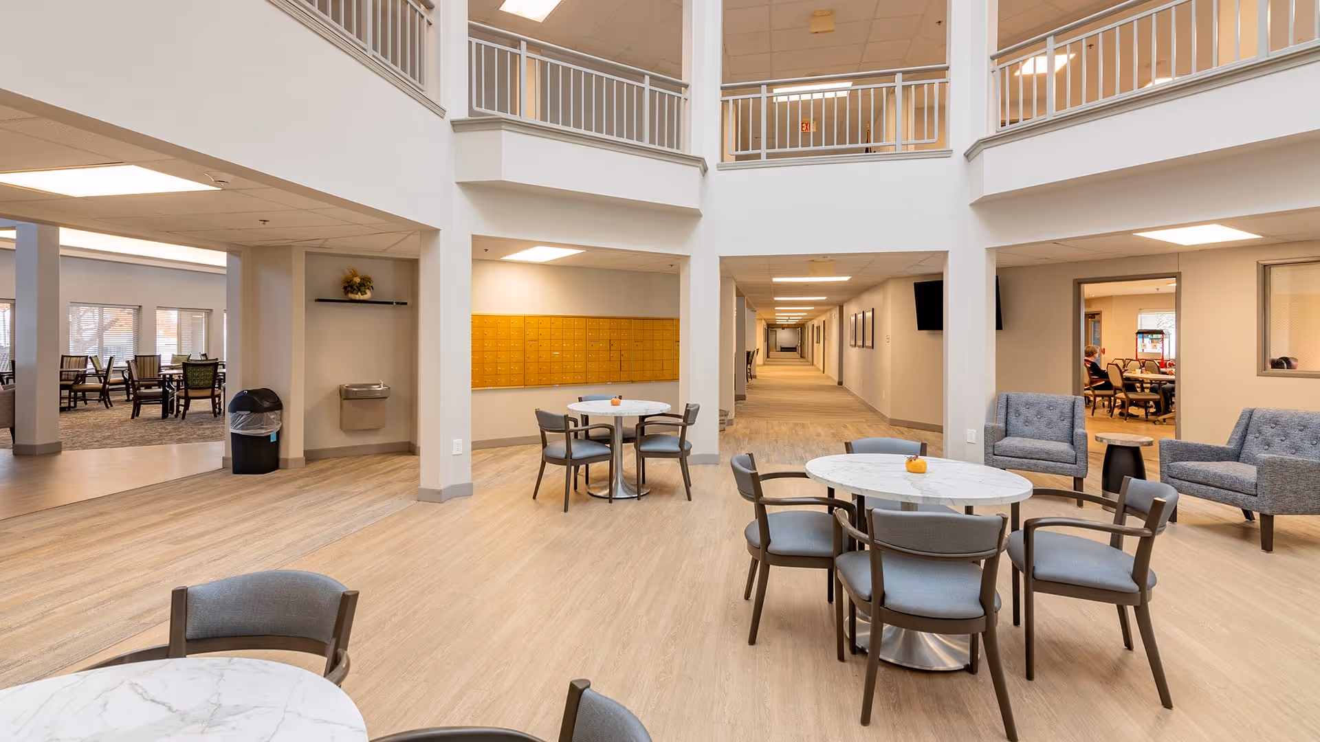 A spacious, well-lit common area in a senior living facility with round tables and chairs arranged for socializing. There are comfortable armchairs on the right side and a long hallway in the background. The area features light wood flooring, white walls, and a high ceiling with a balcony railing above. A water fountain and a trash bin are visible on the left side, and a wall of mailboxes is mounted on the far wall.