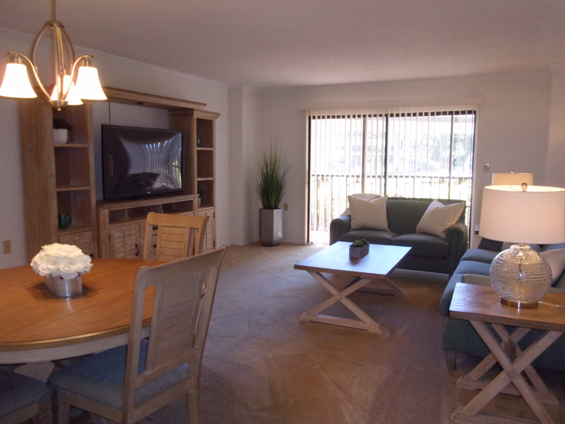 Open living room and dining area with sofas, coffee table, wooden dining table and chairs, TV cabinet, and a large sliding glass door with vertical blinds.