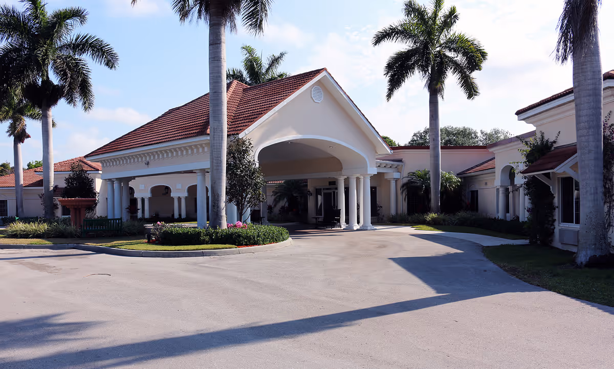 Exterior view of a senior living facility with a covered entrance supported by white columns, surrounded by palm trees and landscaping under a clear sky.
