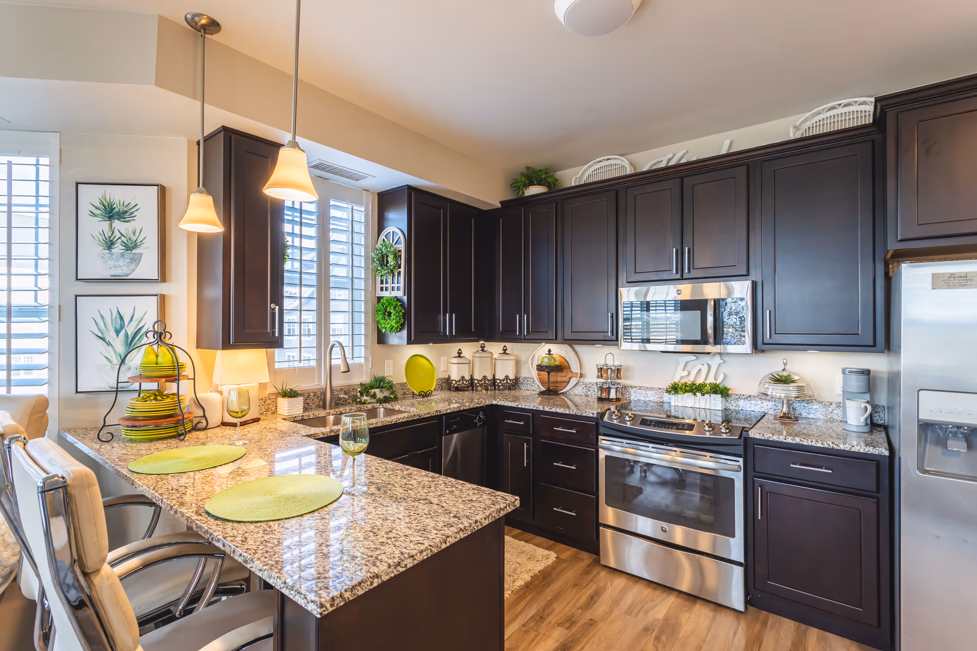 A modern kitchen with dark wood cabinets, granite countertops, and stainless steel appliances including a refrigerator, oven, and microwave. The kitchen features a breakfast bar with two cushioned chairs, green placemats, and wine glasses. There are two pendant lights hanging above the bar, decorative plants, and framed botanical prints on the wall near a window with white shutters.