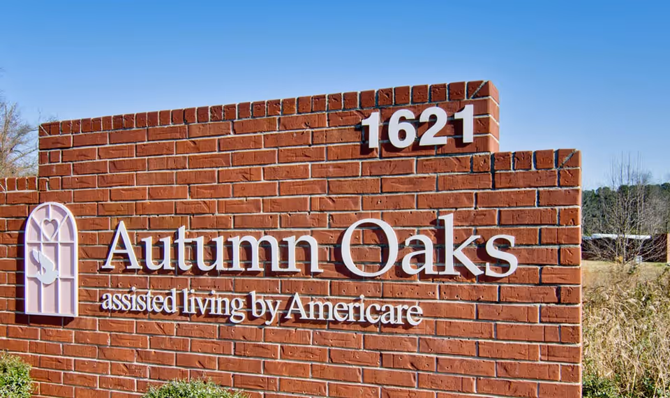 A brick sign for Autumn Oaks assisted living by Americare with the street number 1621 displayed on top, set against a clear blue sky and some greenery.