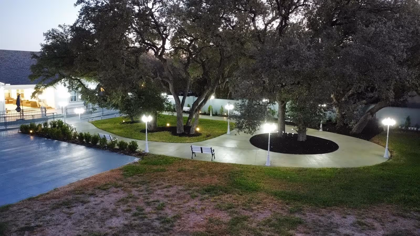 Outdoor garden area at dusk with large trees, paved walkways, white benches, and white lamp posts providing illumination. A building with bright interior lights is visible in the background.