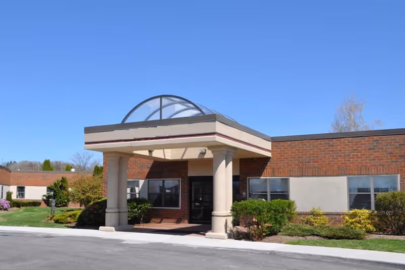 Exterior front view of a single-story brick building with a covered entrance supported by two columns and a glass dome roof feature, surrounded by green shrubs and a clear blue sky.