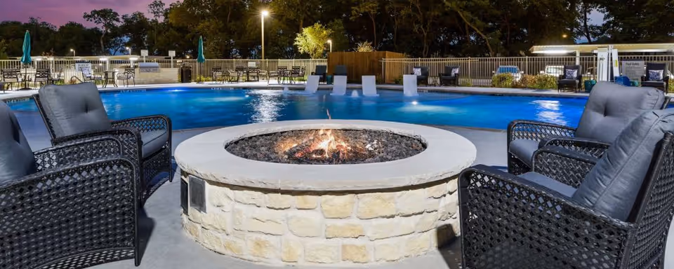 Outdoor pool area at dusk with a central round stone fire pit surrounded by cushioned wicker chairs and lounge seating.