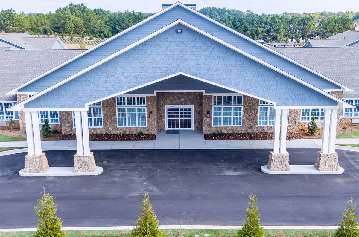 Front exterior view of Manor Lake Assisted Living & Memory Care - Hiram showing a large covered entrance with stone pillars, blue siding, multiple windows, and a paved driveway with greenery in the foreground.