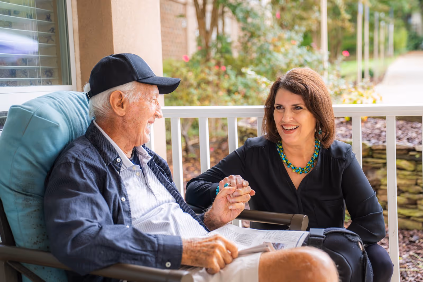 An elderly man wearing a black cap and a woman with a turquoise necklace are sitting and holding hands on a porch with a garden in the background, both smiling and engaged in conversation.