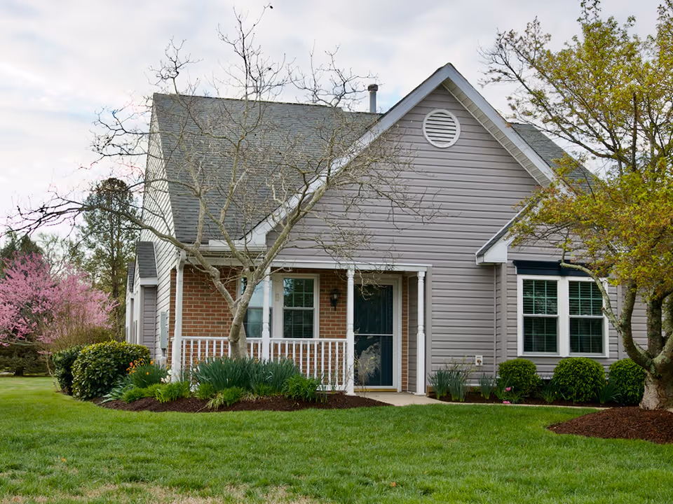 Exterior view of a single-story residential building with gray siding and a brick front porch. The porch has white railings and a dark front door. There are leafless trees and green shrubs in the landscaped yard with a well-maintained lawn. The sky is partly cloudy.
