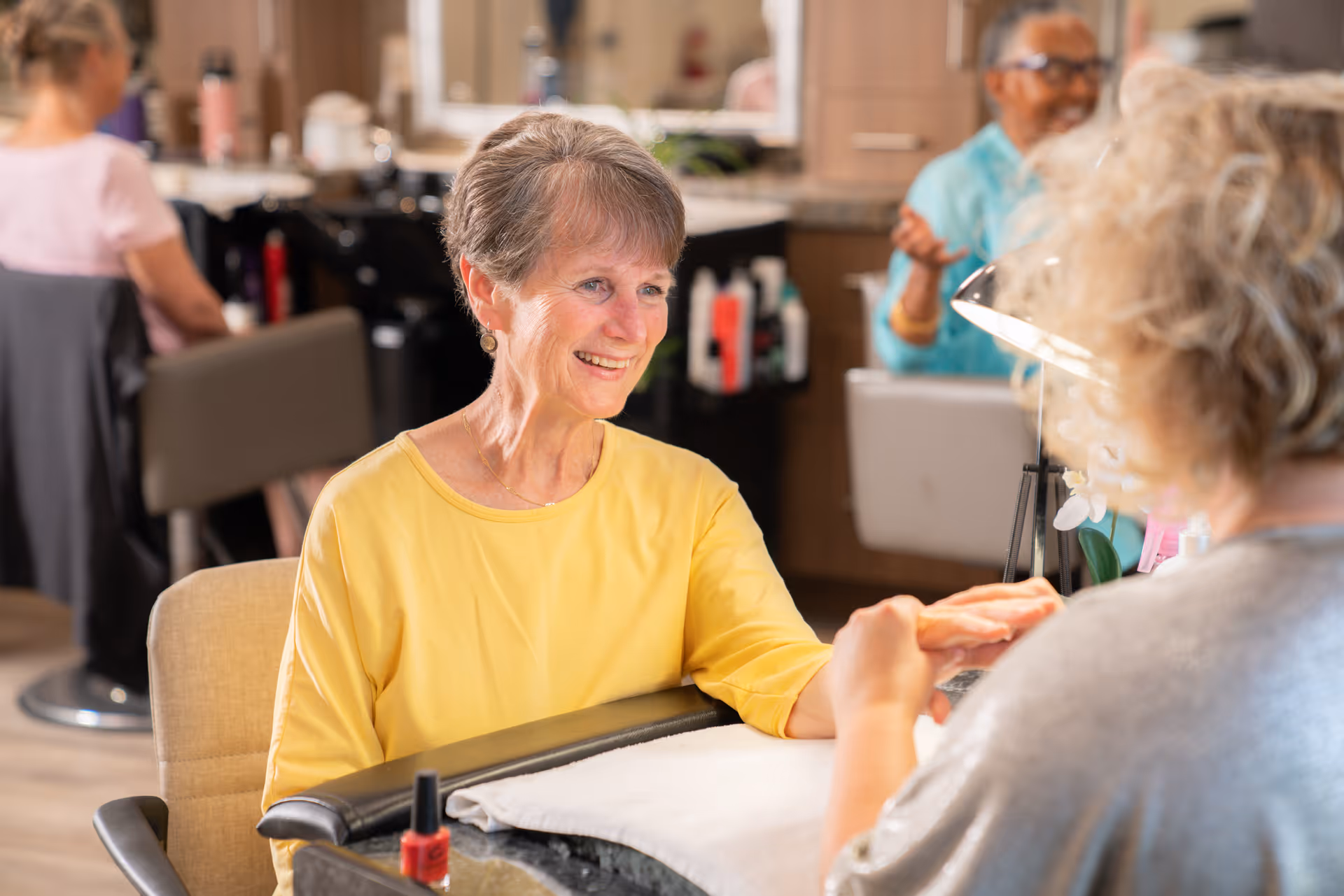 An elderly woman in a yellow shirt smiling while getting a manicure at a salon area inside a senior living facility. Another elderly woman is visible in the background receiving a similar service.