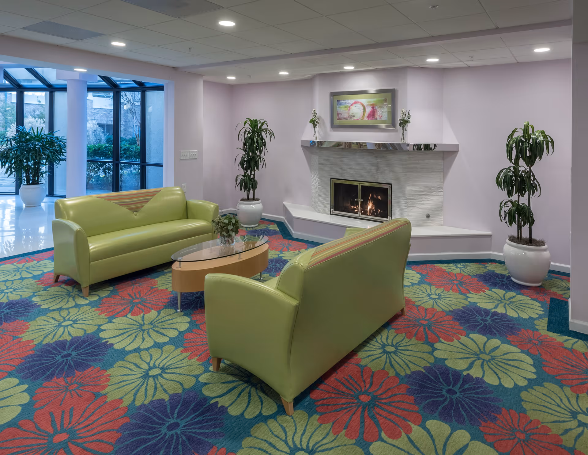 A senior living facility lounge area with two green sofas facing each other around a glass coffee table on a colorful floral carpet. There is a fireplace with a modern white stone surround and a framed artwork above it. Potted plants are placed around the room, and large windows let in natural light.