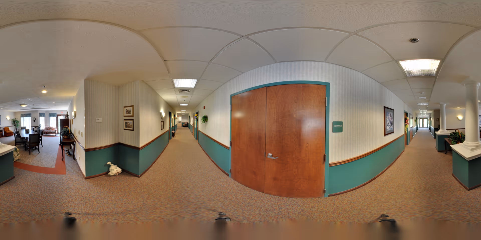 A wide hallway in a senior living facility with beige and teal walls, carpeted floor, and ceiling lights. On the left side, there is a seating area with chairs and a table near windows. A wooden door labeled 'Restroom' is visible on the right side of the hallway. The hallway extends into the distance with framed pictures and plants along the walls.
