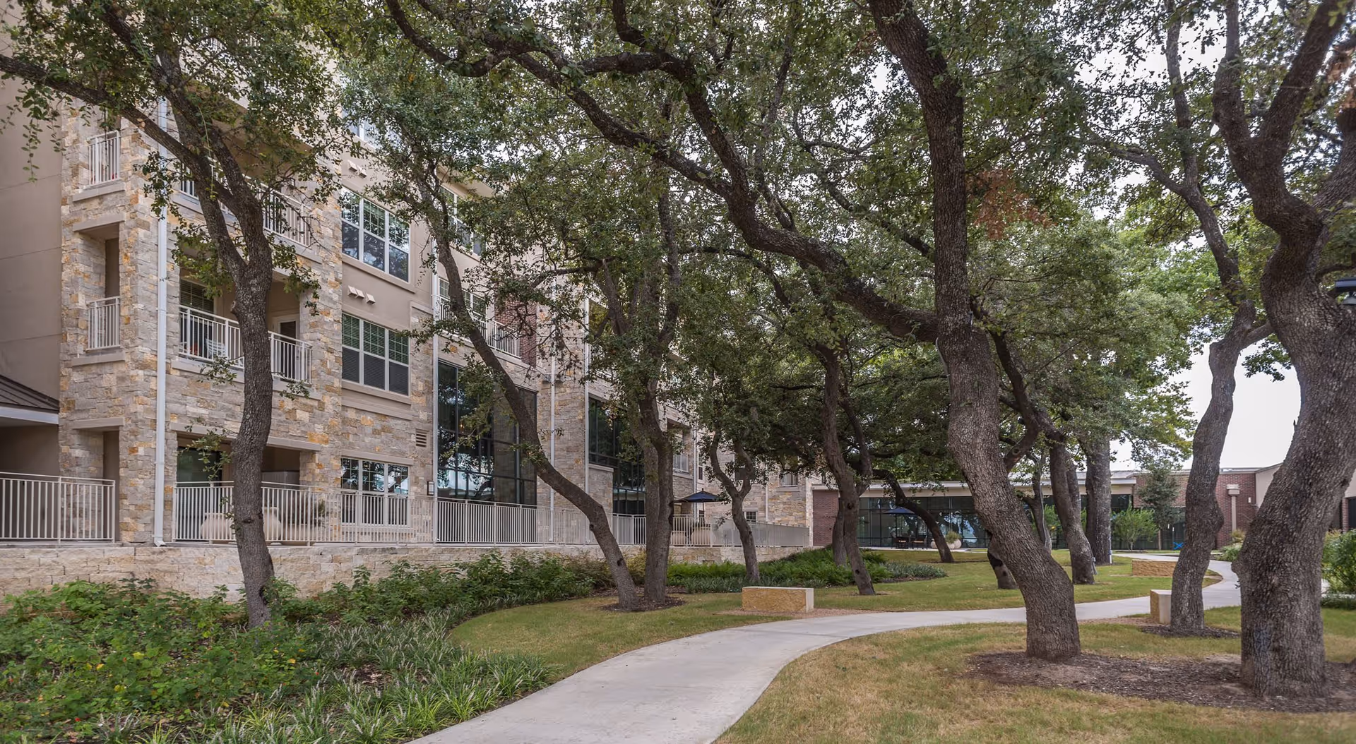 Outdoor view of a senior living facility with a stone and stucco building on the left, surrounded by mature trees and a winding concrete pathway through a landscaped garden area with grass and shrubs.