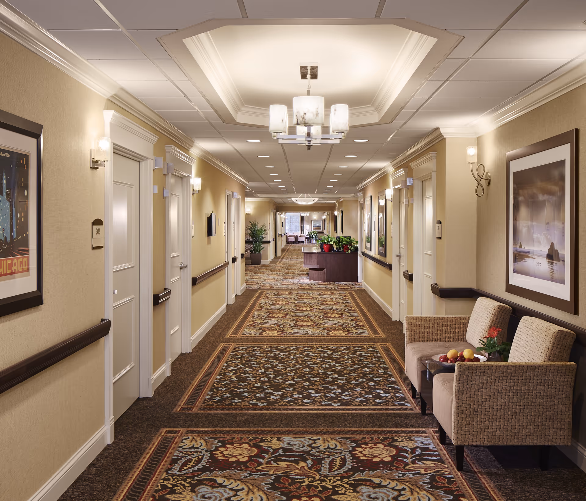 Carpeted, well-lit interior corridor of a senior living facility with chairs and a small table on the right, doors and framed artwork along the walls, and decorative ceiling lights.