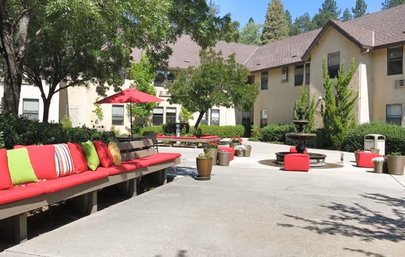 Sunny outdoor courtyard with red-cushioned benches, potted plants, a central fountain, and surrounding two-story buildings.