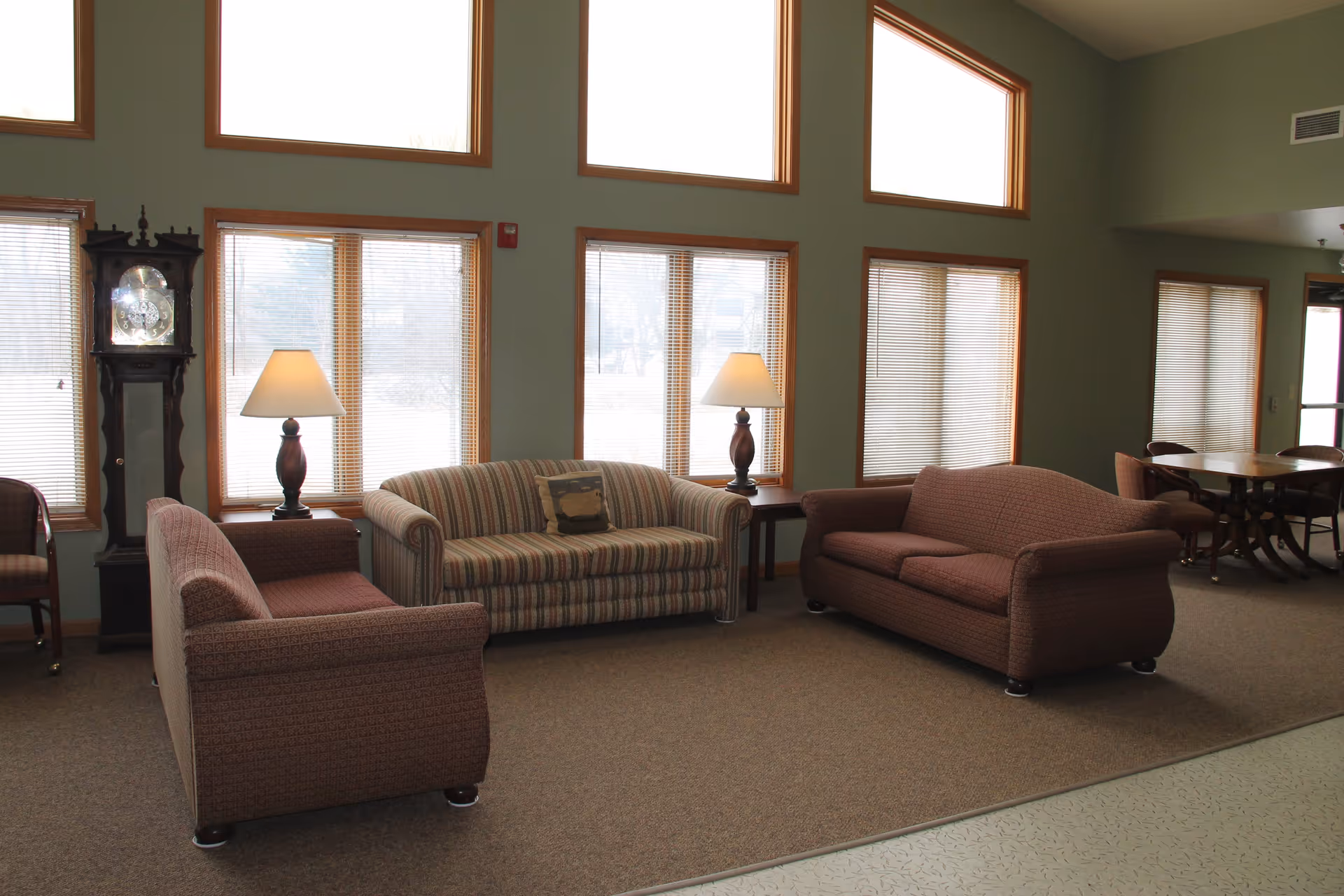 Communal living room with three sofas, two table lamps, large windows, and a grandfather clock.