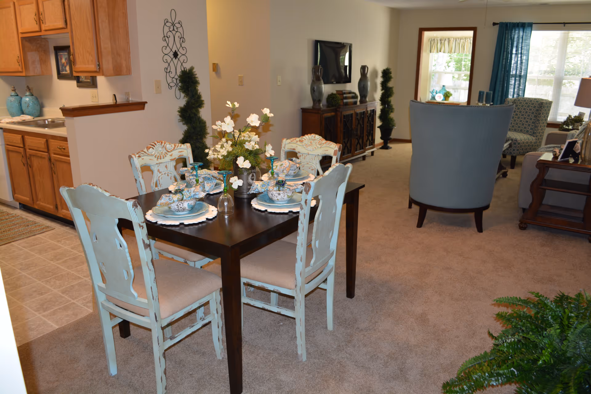 A cozy interior space featuring a dining area with a dark wooden table set for four with vintage-style chairs painted in a distressed light blue. The table is decorated with floral arrangements and tableware. To the left is a kitchen area with wooden cabinets and a tiled floor. In the background, there is a living room area with upholstered chairs, a TV on a wooden cabinet, and large windows letting in natural light.