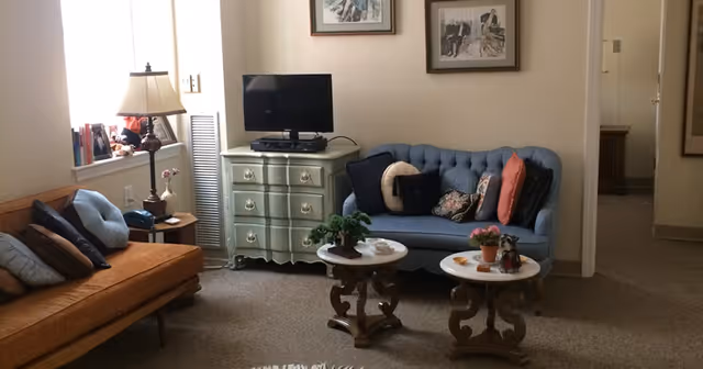 A cozy living room with a blue tufted sofa adorned with multiple pillows, an orange cushioned bench with pillows, a small green dresser with a TV on top, two round wooden side tables with plants and decorative items, a floor lamp, and framed artwork on the walls.