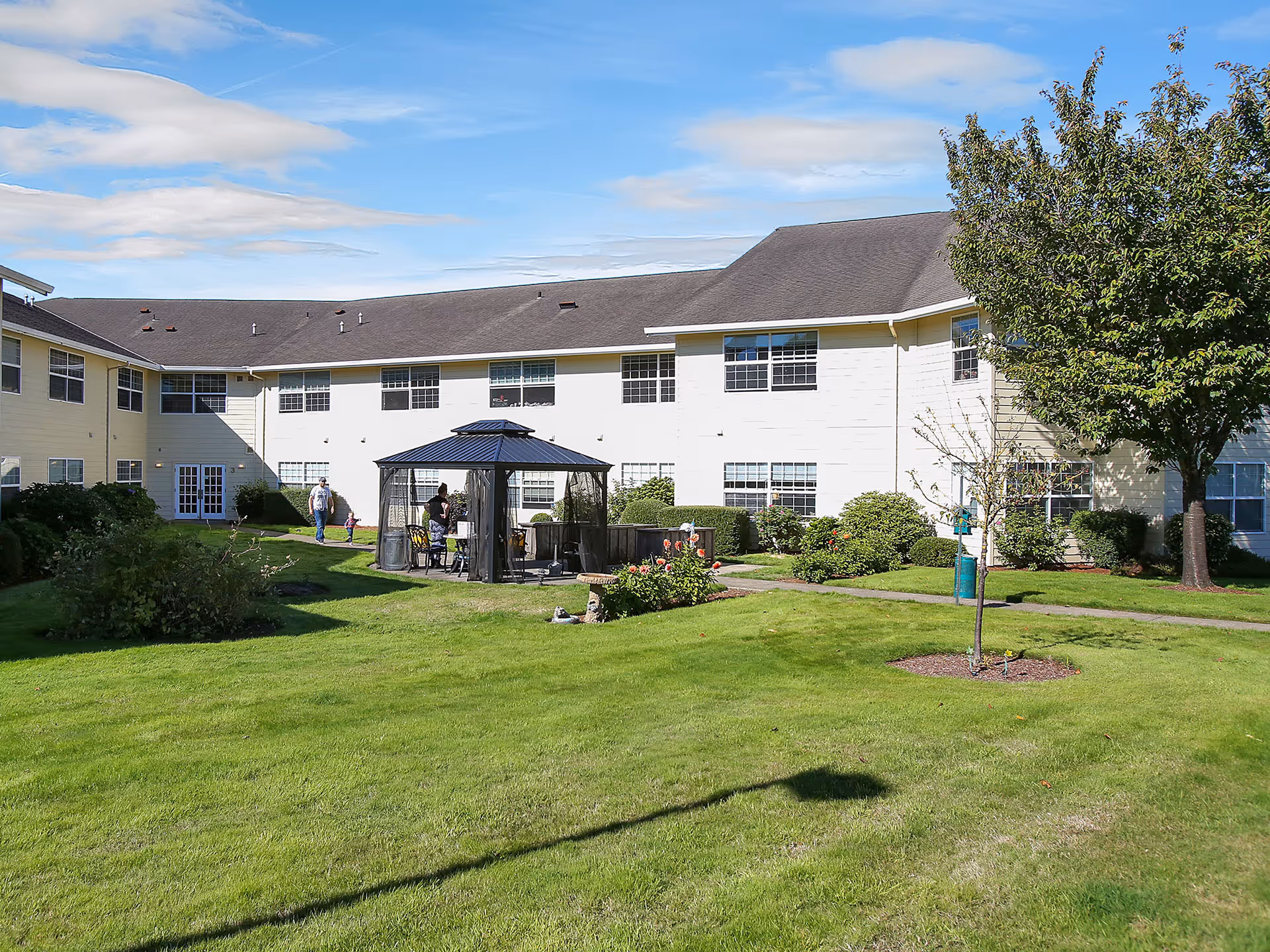 Well-maintained senior living courtyard with a gazebo, green lawn, shrubs, and a two-story residential building in the background.