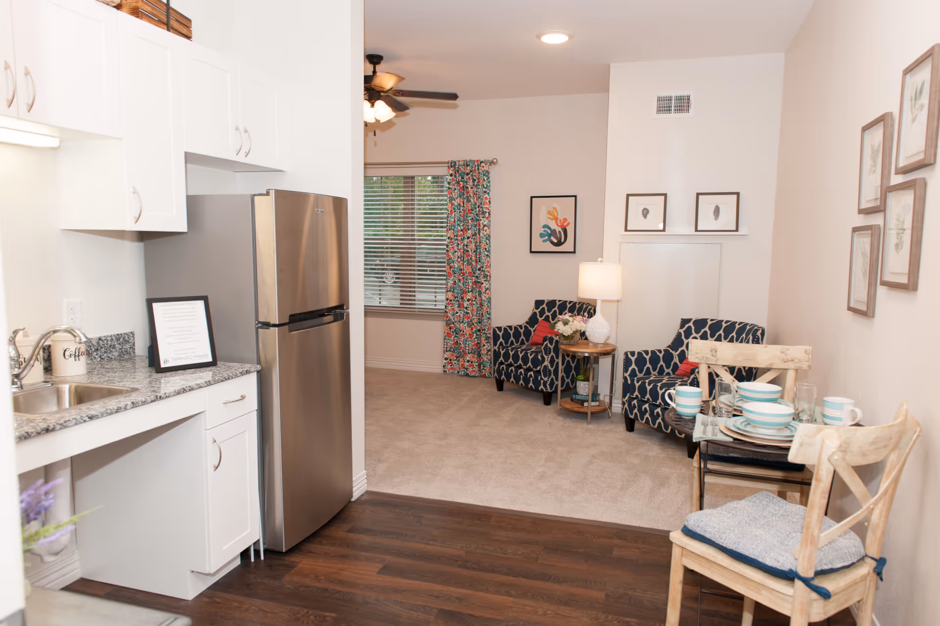 Open-plan apartment interior with a kitchenette and stainless refrigerator leading into a cozy living area with patterned armchairs, a small dining table, and decorative accents.