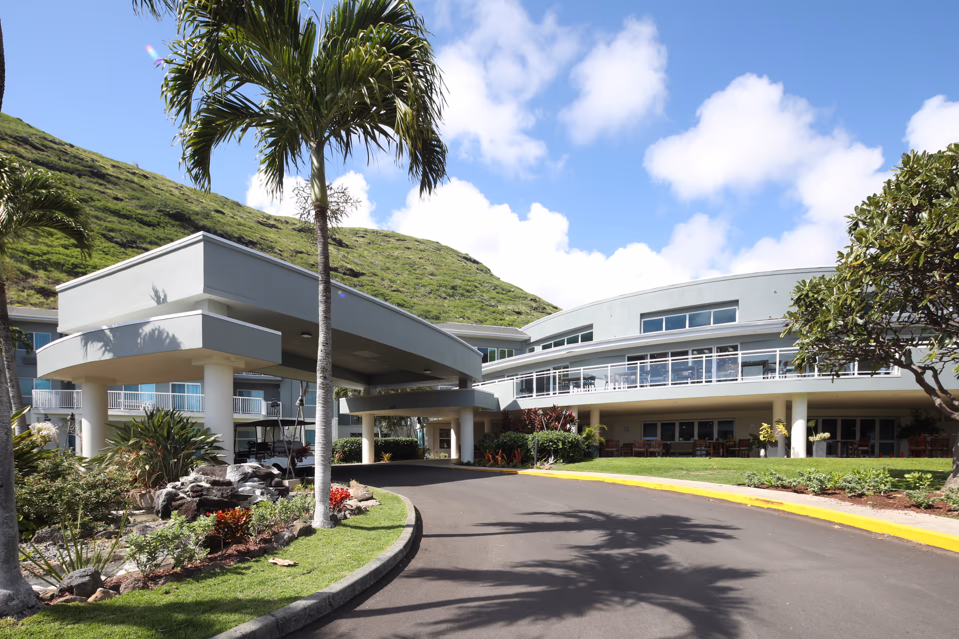 Exterior view of The Ivy at Hawaii Kai senior living facility with a covered driveway entrance, palm trees, landscaped garden, and a green hillside in the background under a partly cloudy sky.