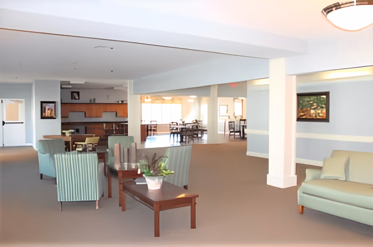 Spacious senior living common area with sofas, armchairs, coffee tables, and a dining area visible in the background.