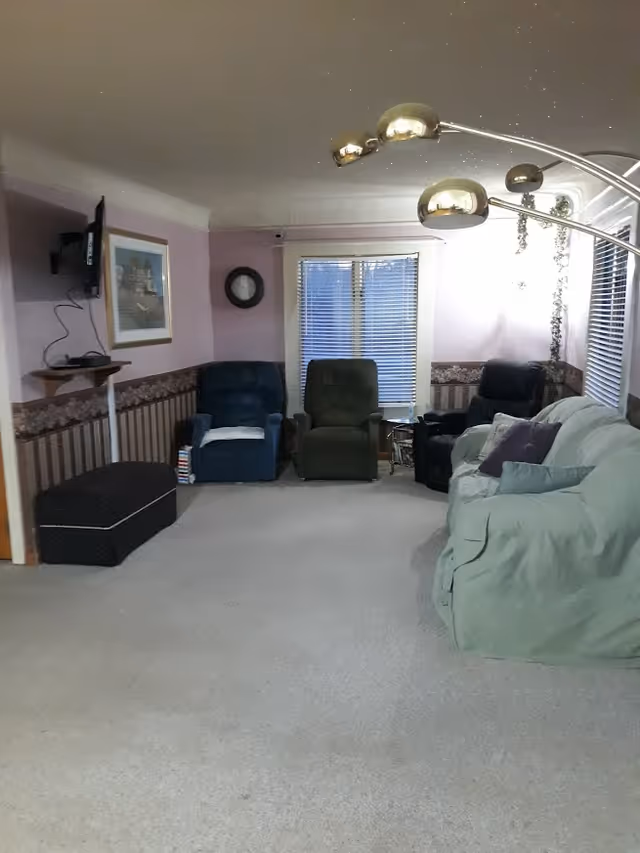 A cozy living room with several recliner chairs and a covered sofa facing a wall-mounted TV and window with blinds.