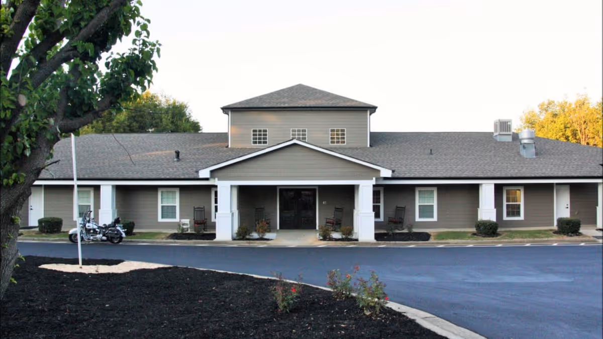 Front exterior of a single-story senior living building with a covered entrance, white columns, rocking chairs, and a paved driveway.