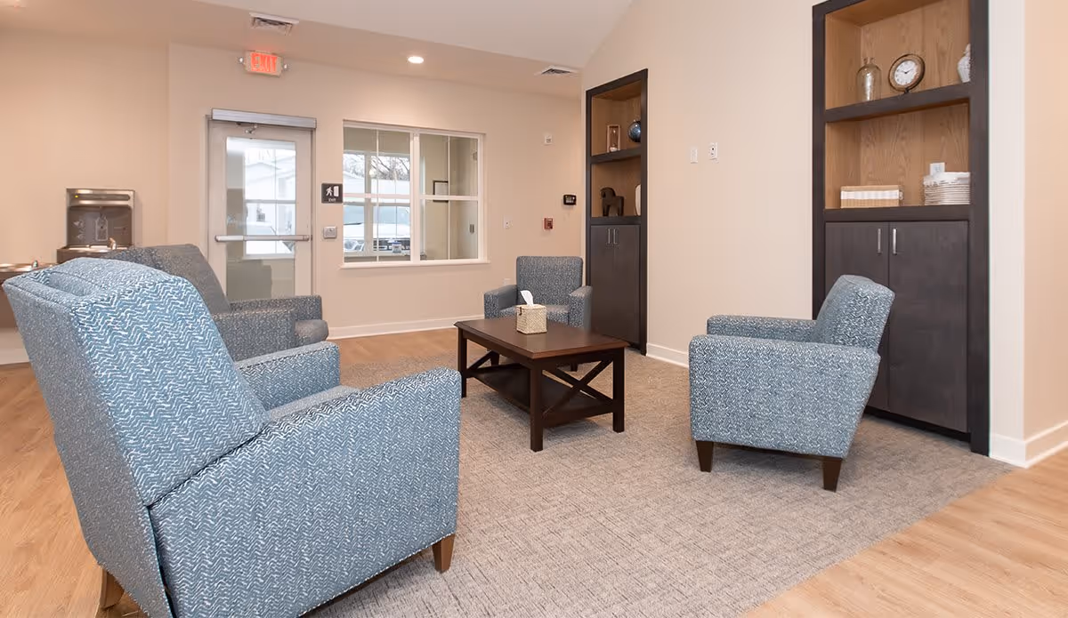 A cozy sitting area in an assisted living facility featuring four blue upholstered armchairs arranged around a dark wooden coffee table with a tissue box on top. The room has light beige walls, a carpeted floor area under the chairs, and wooden flooring around it. There are built-in shelves with decorative items and plates on the right wall, a window and an exit door in the background, and a water dispenser on the left side.
