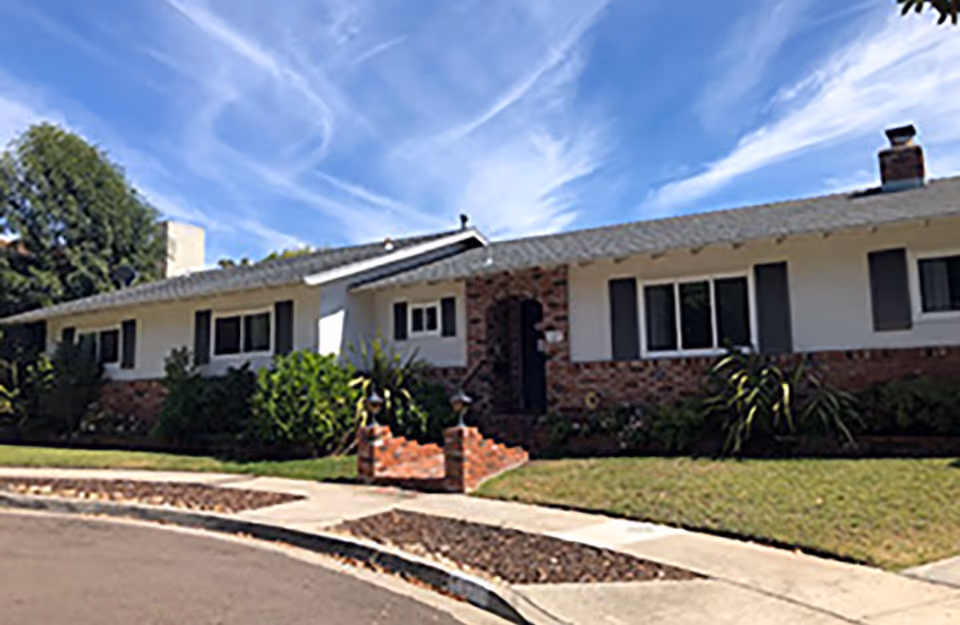 Single-story brick-and-stucco ranch-style house with a front lawn, walkway, and shrubs under a blue sky.