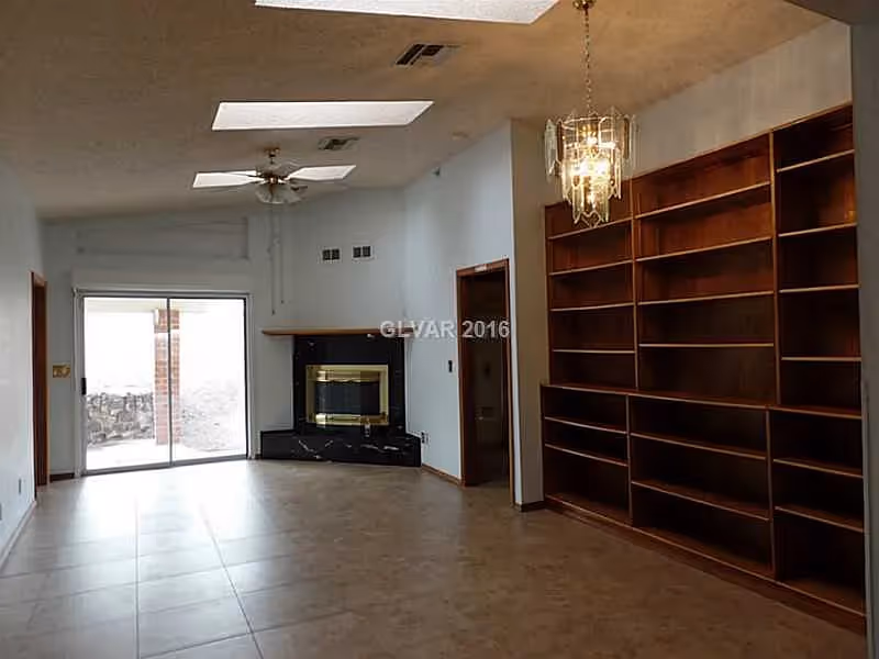 A spacious living room with tiled flooring, a ceiling fan, and a chandelier. There is a large built-in wooden bookshelf on the right wall and a fireplace with a black marble surround at the far end. Sliding glass doors lead to an outdoor area, and there are skylights in the ceiling providing natural light.