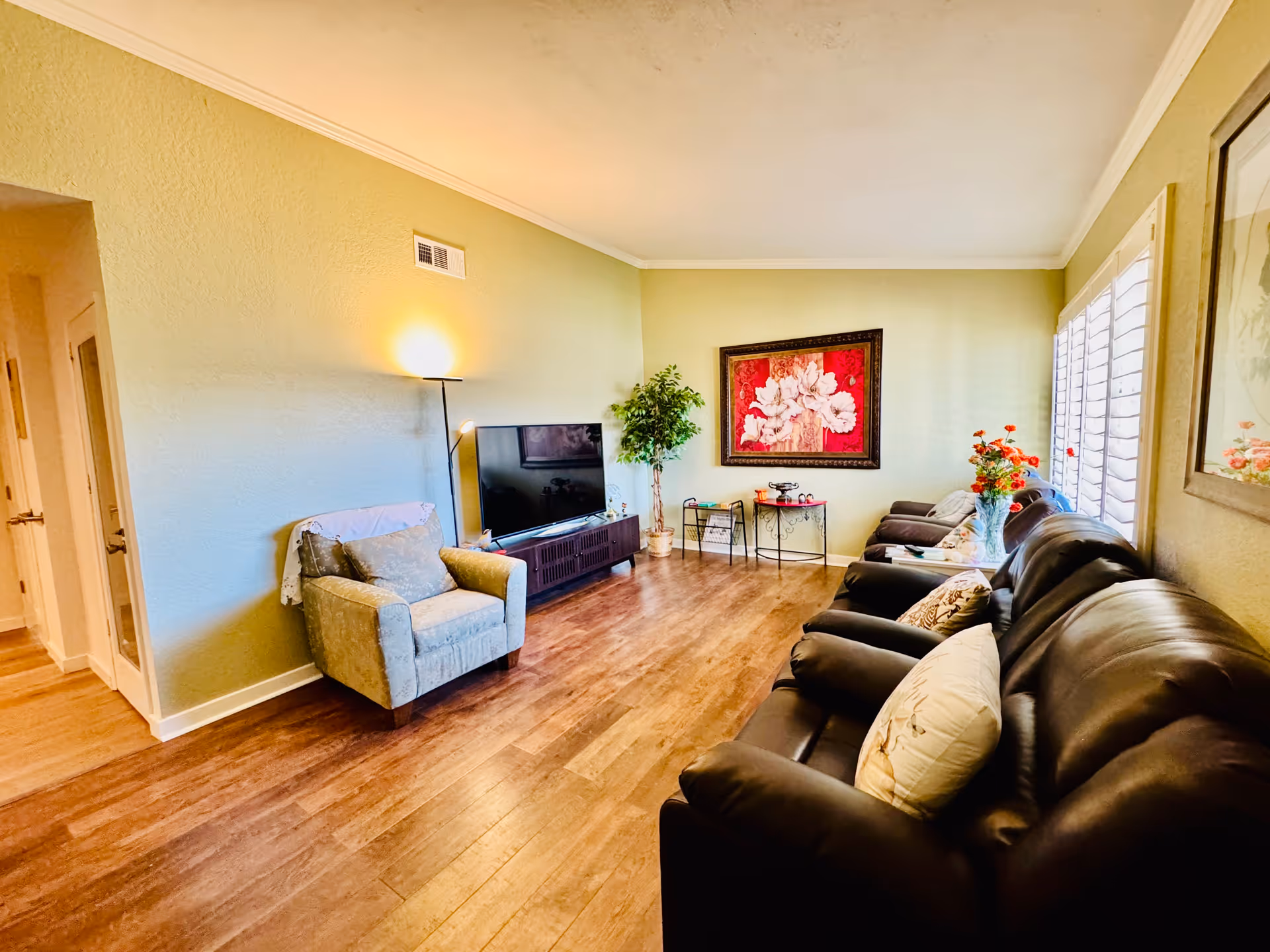 A cozy living room with wooden flooring, a large black leather sofa with pillows, a single armchair with a throw blanket, a flat-screen TV on a stand, a floor lamp, a potted plant, a small table with decorative items, and a large framed floral artwork on the wall. Sunlight streams in through white plantation shutters on the window.