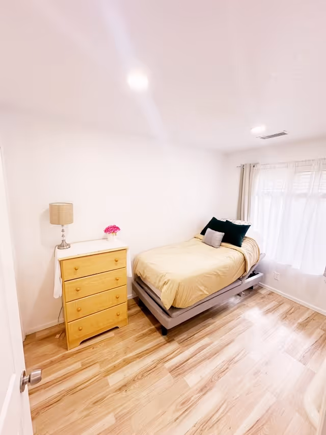 A small, bright bedroom with light wood flooring, a single bed with beige bedding and multiple pillows, a wooden dresser with a lamp and a small flower vase on top, and a window with sheer white curtains allowing natural light to fill the room.