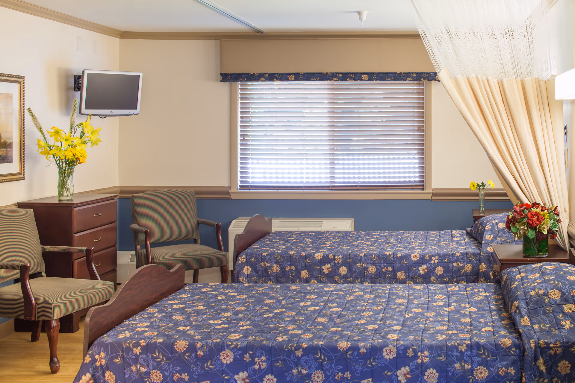 A well-lit bedroom with two twin beds covered in blue floral bedspreads. There are two upholstered chairs, a wooden dresser with a vase of yellow flowers, a small wall-mounted TV, and a window with closed blinds. A curtain is partially drawn on the right side, and a nightstand with a vase of red and yellow flowers is between the beds.