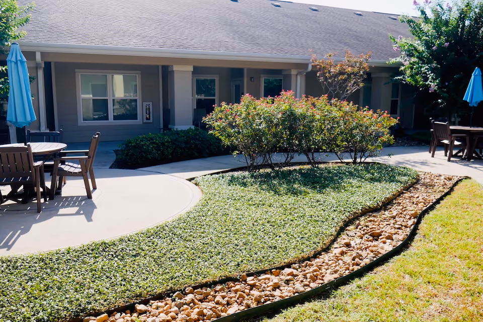 Outdoor patio area at Orchard Park at Victory Lakes Assisted & Senior Living featuring round tables with chairs and blue umbrellas, surrounded by landscaped greenery, bushes, and a rock-lined garden bed in front of a single-story building.