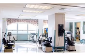 Bright and spacious rehabilitation room with exercise equipment including stationary bikes and other physical therapy machines, large windows with floral valances allowing natural light, and a clean, polished floor.