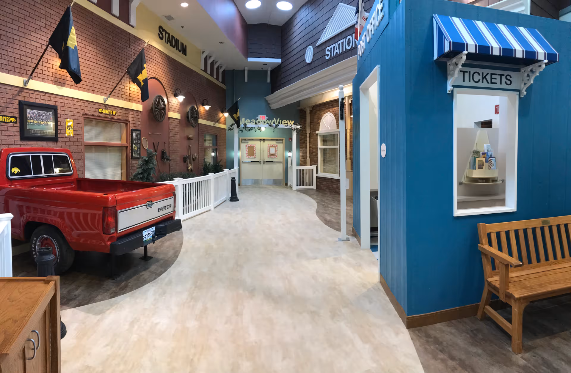 Indoor hallway designed to resemble an outdoor street scene with a red vintage pickup truck, brick walls with black and yellow flags, a blue ticket booth with a striped awning, wooden bench, and various storefront facades including one labeled 'STADIUM' and another 'STATION'.