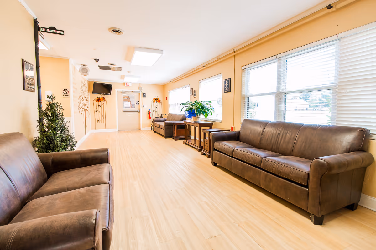 A bright and clean common area with light wood flooring and beige walls. The room features two brown leather sofas facing each other along the walls, a small table with a green plant, and large windows with white blinds letting in natural light. There is a TV mounted on the far wall, a fire extinguisher, and decorative wall art near the exit door.