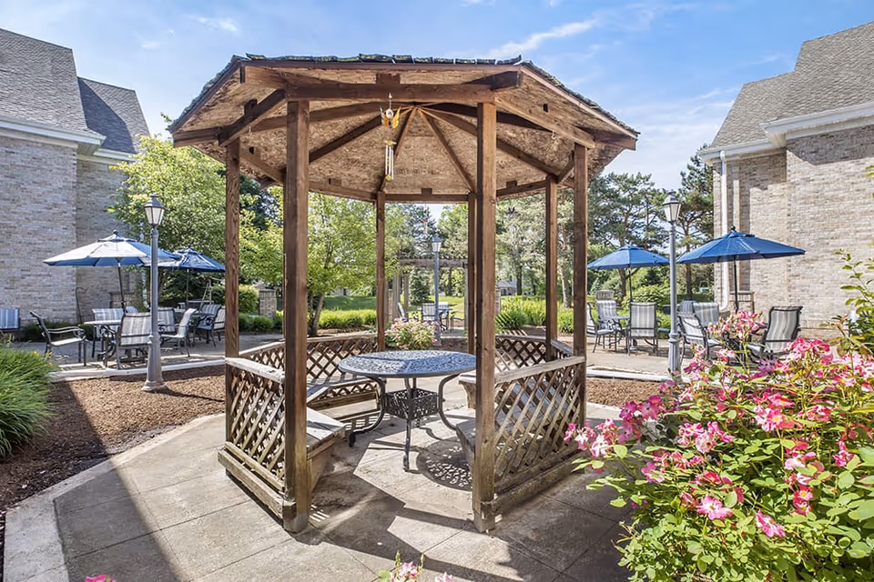 Outdoor seating area at a senior living facility featuring a wooden gazebo with a round metal table and benches inside. Surrounding the gazebo are patio tables with blue umbrellas and chairs, set on a paved area with greenery and pink flowers nearby. The background includes brick buildings and trees under a clear blue sky.