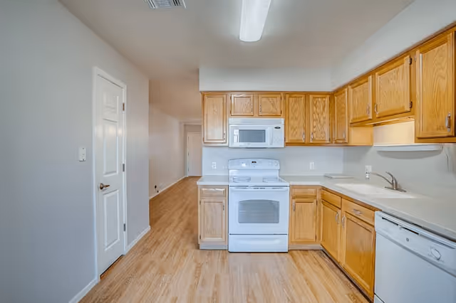 A clean kitchen with wooden cabinets, white countertops, and white appliances including a stove, microwave, and dishwasher. The kitchen has light wood flooring and a white door on the left side leading to another room. The space is well-lit with a fluorescent ceiling light.