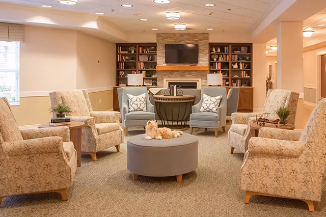 Cozy senior living lounge with upholstered armchairs arranged around a round ottoman facing a fireplace, TV, and built-in bookshelves.