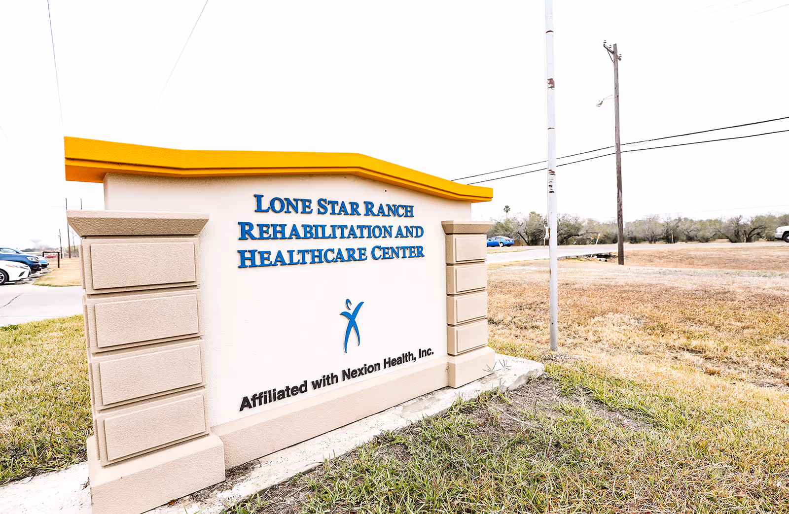 Outdoor sign for Lone Star Ranch Rehabilitation and Healthcare Center with a yellow top and beige pillars on each side, located on a grassy area near a road with some cars and utility poles in the background.