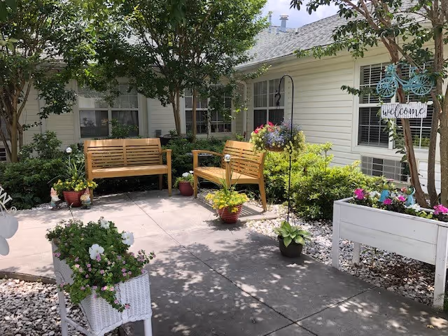 Outdoor garden area with two wooden benches surrounded by various potted plants and flowers. The area is paved with concrete and bordered by white gravel. There is a white planter box on the right side with colorful flowers and a decorative sign that says 'Welcome'. Trees and shrubs provide shade and greenery around the seating area, adjacent to a light-colored building with windows.