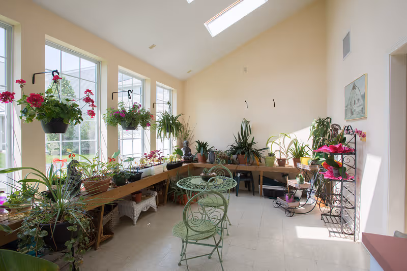A bright sunroom with large windows and skylights, filled with various potted plants on wooden shelves and hanging baskets. In the center, there is a small green metal table with two matching chairs. The room has light-colored walls and floor, and a painting hangs on the right wall.
