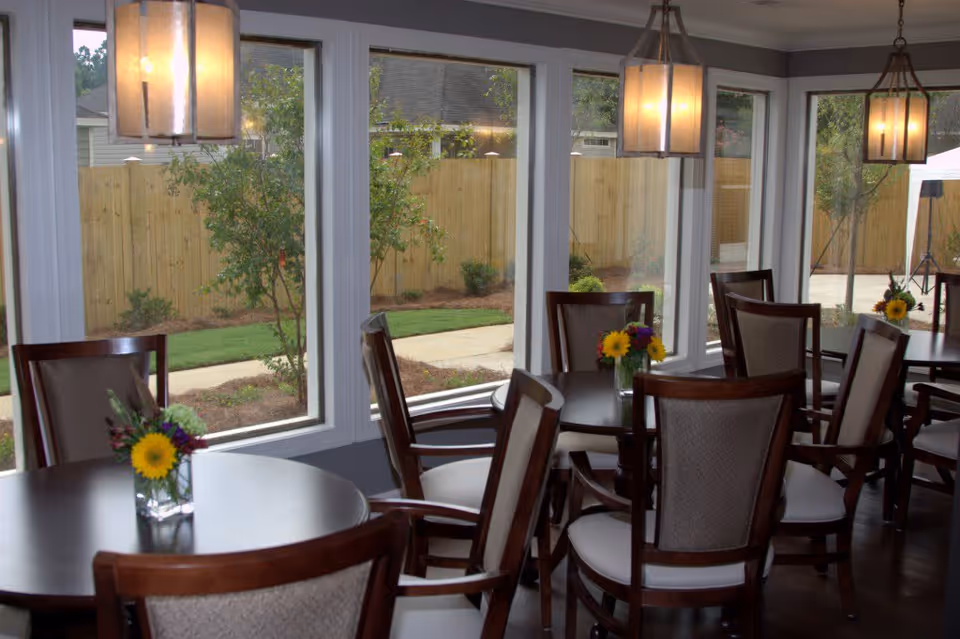 Dining area with round wooden tables and cushioned chairs, each table decorated with a small vase of flowers. Large windows provide a view of a fenced garden with a walkway and greenery outside. Pendant lights hang from the ceiling, illuminating the space.