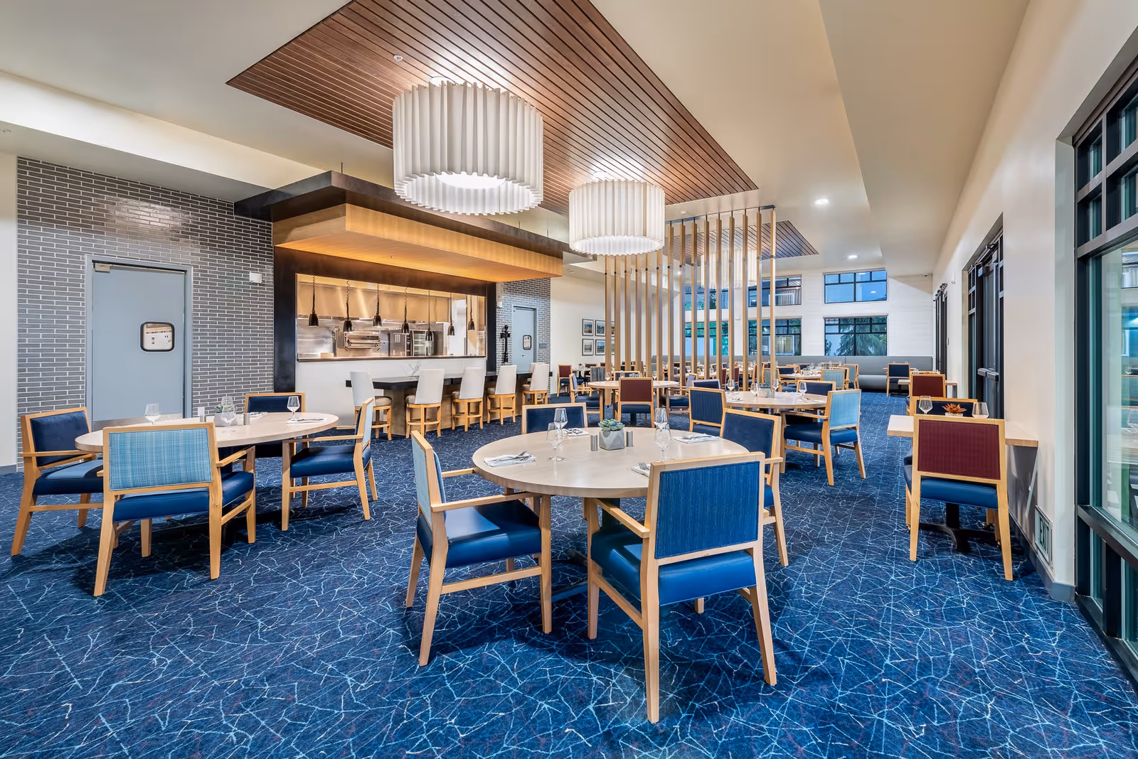 A spacious dining room with round and rectangular tables surrounded by wooden chairs with blue and red upholstery. The room features a blue patterned carpet, large windows on the right side, and modern pendant lights hanging from a wooden ceiling panel. In the background, there is a kitchen area with a counter and bar stools.