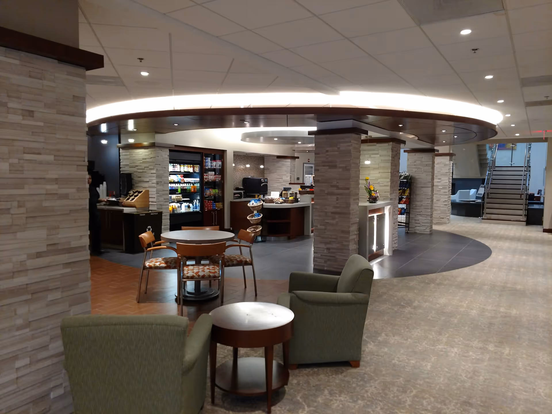 Interior view of a senior living facility common area featuring a circular counter with snacks and beverages, surrounded by stone pillars. There are tables and chairs for seating, two green armchairs with a small round table in the foreground, and a staircase in the background.