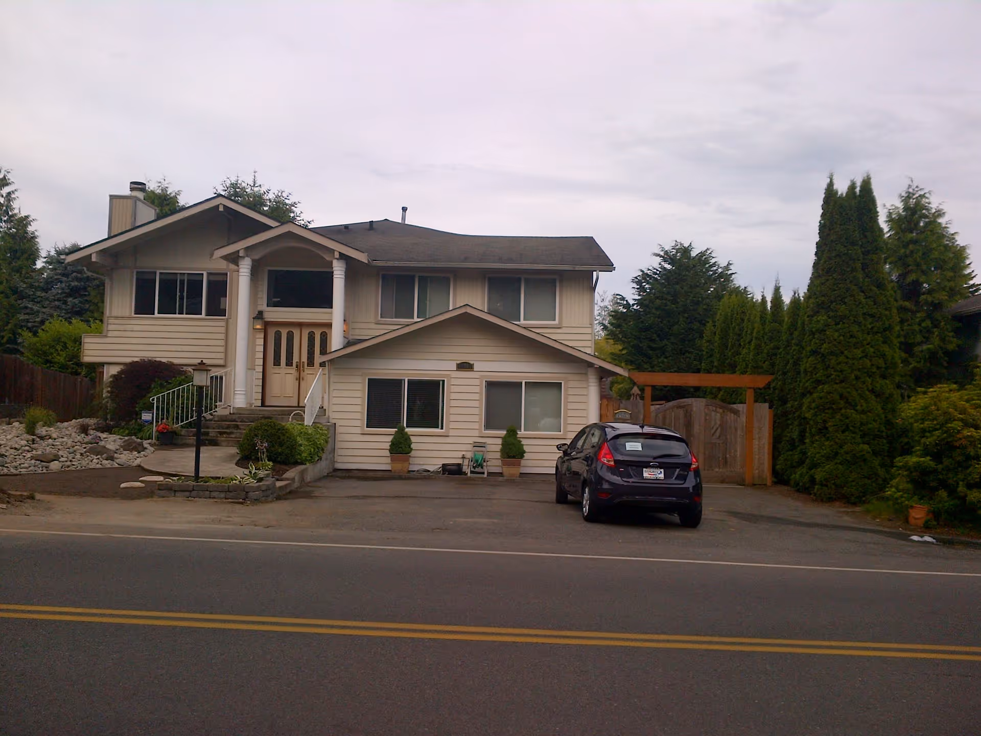 Front exterior view of a two-story beige house with white columns at the entrance, a driveway with a parked black car, and surrounding greenery including trees and shrubs.