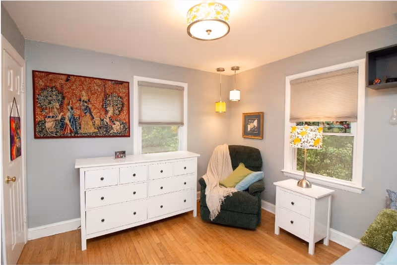 A cozy room with light gray walls and wooden floor featuring a white dresser with multiple drawers, a green armchair with a white throw blanket and colorful cushions, two windows with beige blinds, a white nightstand with a floral lampshade, and decorative wall art including a tapestry and a framed picture.